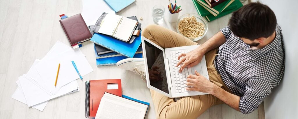 a man sitting on the ground and working on a laptop, surrounded by notebooks