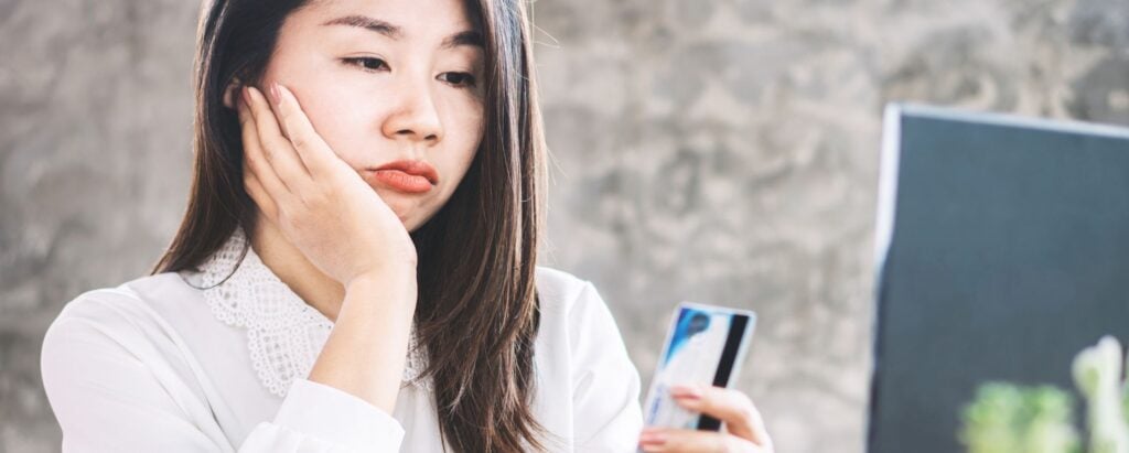 A woman is looking worryingly at the bank card in her hand in front of the computer